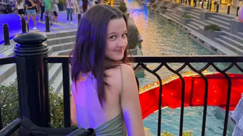 Maddie Haining Maddie Haining smiles as she wear a dress while sitting looking out over a pool