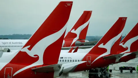 Four planes are lined up in a row on the tarmac of an aiport, each with the Qantas logo of a stylized white kangaroo on a red background emblazoned on the tail. 
