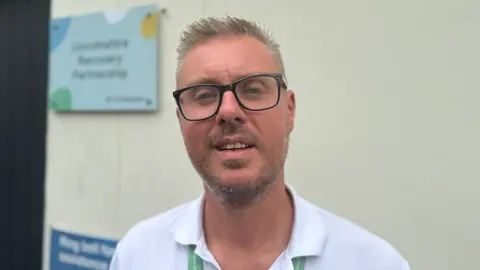 BBC The photo shows a man looking at the camera. He's got short, spiky blonde hair. He's wearing black frame glasses, and a white top with a colour. He's standing in front of a white building.