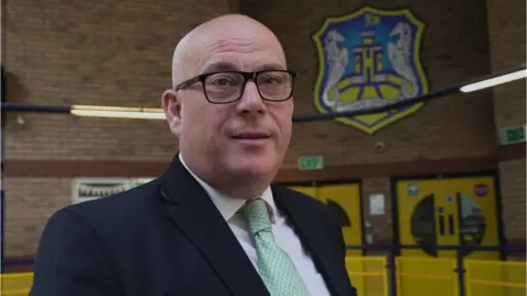 Gemma Laister Man standing in a school wearing glasses looks at camera