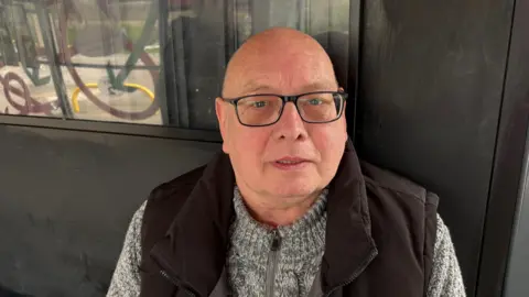 Martin Heath/BBC Gordon Boyd with very short hair and glasses, looking at the camera and wearing a brown gilet over a grey sweater. He is sitting in a bus shelter.