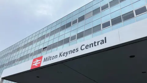 The outside of Milton Keynes railway station. There is a sign with the station name and glass windows above.