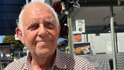 James Moule is wearing a patterned short‑sleeve shirt, with shops and a bus shelter visible behind him.