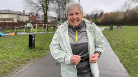 Barbara stands on a paved path in an outdoor park setting. She is wearing a light green jacket over a grey athletic top with a yellow zipper detail. In the background, there is a grassy area with scattered trees, a few green bins, and some rope barriers marking off sections of the park. Residential houses are visible further back, along with a lamppost.