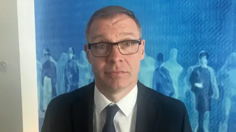 A man with glasses and a dark suit, white shirt and navy tie on. 