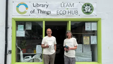 A man and a woman stand outside a shop with yellow painted windows while holding a heat gun and a electric mixer. Above the door is a sign which reads Library of Things and Leam Eco hub.