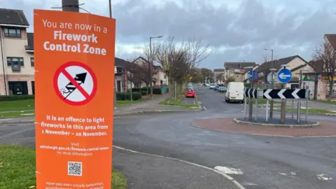 BBC Orange sing tied to a lamp post saying firework control zone just in front of a roundabout with some houses in background