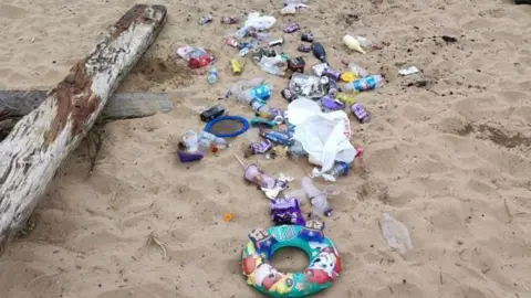 Durham Wildlife Trust Child's rubber beach ring, plastic bottles, paper strewn on a beach beside a log of wood. 