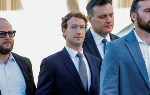 Meta boss Mark Zuckerberg arrives at an LA court wearing a dark blue suit jacket, white shirt and grey tie. He is surrounded by security men
