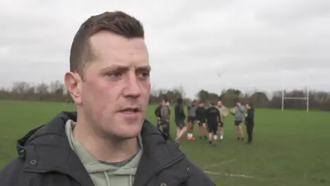Chris is standing to the side of the pitch, with a training session taking place behind him. He is looking off camera, the sky behind him is grey.