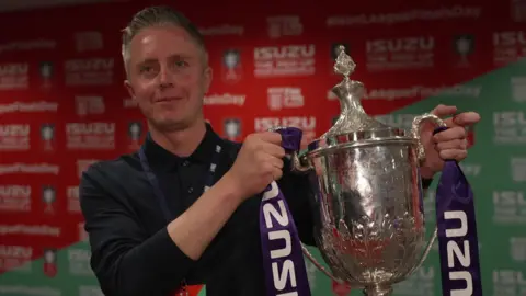 James Phillips smiles as he holds the FA Vase - a large silver trophy - in a room. He wears a black shirt buttoned up to the neck and has combed-back greying hair.