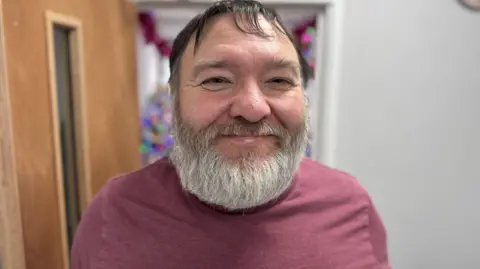 A man with a pink shirt smiles and looks into the camera.