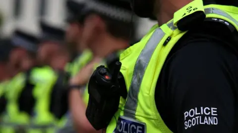 A close up of a line of police offices in black Police Scotland branded jumpers and very bright yellow-green visibility vests. We can see a police radio in the foreground and the rest of the picture beyond the first officer is out of focus. We can see no faces.