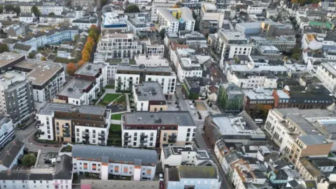 BBC A drone shot of buildings and homes in St Helier in Jersey. The photo shows roof tops and a number of roads and trees between the buildings.