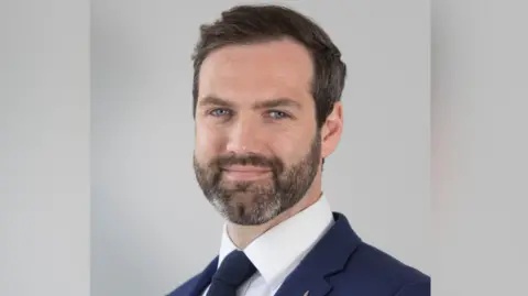 RenewablesNI A headshot of Mark Richardson. He is smiling at the camera and is placed in front of a grey background. He is a middle-aged man, with dark, short hair and a beard with some grey patches. He is wearing a navy suit and tie, with a white collared shirt.