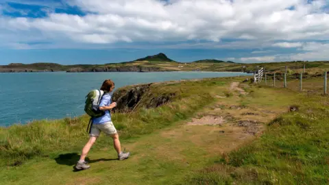 A woman in a blue shirt and cream shorts walking along a coastal path