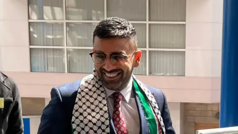 Akhmed Yakoob wearing a suit and glasses. He has short dark hair and is smiling. He has a red tie and is wearing a brown and white patterned scarf as well as a green, white and red one underneath