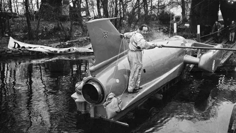 Mirrorpix via Getty Images Donald Campbell, dressed in a pilot's outfit, stands on the side of Bluebird as it is lowered into Coniston Water on a ramp.