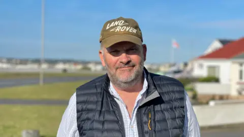 A man wearing a cap is looking towards a camera. He is standing outdoors in a residential area. The sky is blue. 