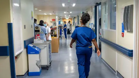 PA Woman in blue uniform walks along hospital corridor - medical staff with mask on are on left of her - doors to rooms line the corridor