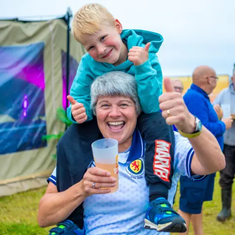 Tiree Music Festival A person carrying a child on their shoulders gives a thumbs-up while holding a drink cup, with other attendees and festival tents visible in the background.