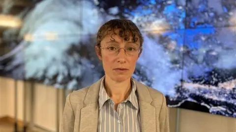 A woman wearing glasses looking towards the camera. She has brown hair tied up and is stood in front of a wall with a storm graphic on it.