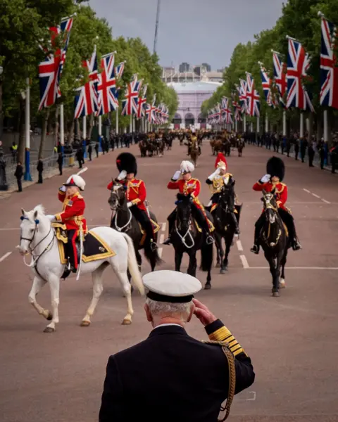 PA Media The King can be seen from the rear as he salutes wearing formal attire and a white military hat, while military personnel on horseback march down the Mall in the background with union flags and trees lining the street.