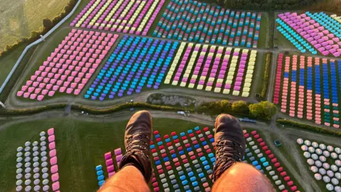 Launch-it Paramotoring Aerial view of Worthy Farm. The paraglider's legs can be seen at the bottom of the frame, and fields are lined with colourful tents