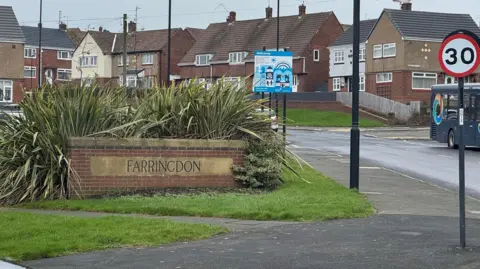 A brick sign on a street saying Farringdon. It is on a patch of grass and there are plants behind it. There are houses in the background.