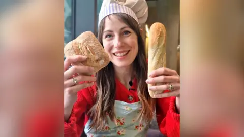 A woman in a red top and chef's hat holding a baguette in one hand and ciabatta in the other