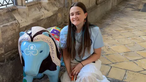 Seren Jaye Seren Jaye kneels in a historic cloister beside a brightly painted elephant sculpture. She is holding a white cane and wear a light top and long white skirt. Sunlight filters through tall leaded windows onto the stone floor and walls.