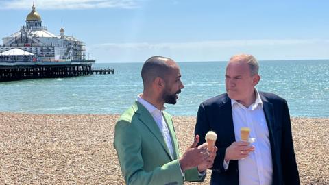 Sir Ed Davey and Josh Babarinde eating ice creams on the beach with Eastbourne Pier in the background