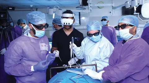 Professor Ajith George Medics take part in training in a simulation course in Delhi at the All India Institute of Medical Sciences. They are wearing head coverings, masks and scrubs and standing around a bed with the equipment. There is operating theatre equipment all around them.