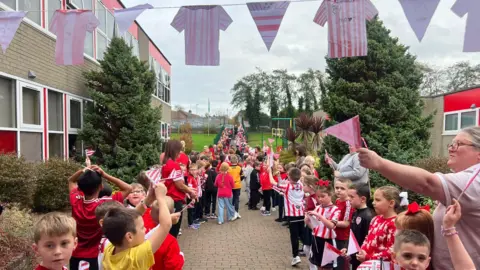 A guard of honour at Steelstown Primary School - pupils wearing red and white Derry City shirts wave flags and there is bunting - which is made up of paper, hand drawn cut outs of Derry shirts