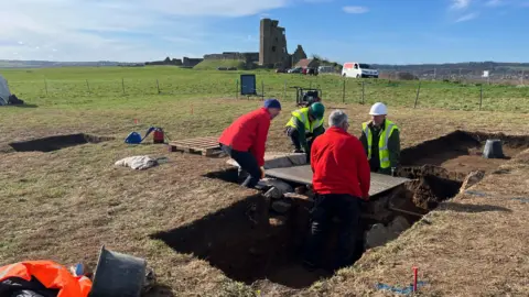 Two people in red coats and two people in high-vix jackets move a metal cover. In the background is Scarborough Castle