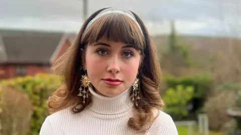 Maddie is stood outside in a back garden. She has light brown hair and fringe. She is wearing a polkadot hair band, white knitted turtleneck jumper, and large gold earrings. She has her nose pierced on the right side of her face and a stud piercing under her bottom lip.