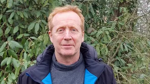 Stefano Biagini looks at the camera, we can see his head and shoulders. He has a grey roll neck jumper on under a dark blue coat. Behind him there are leaves and a tree visible. He has red hair and fair skin.