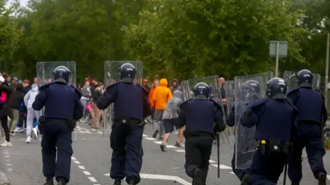 PA Media The Garda Public Order Unit stand in front of protestors near the site of a former factory in Coolock, Dublin, Ireland