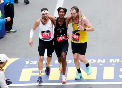 Reuters Three marathon runners move along a finish line with their arms linked over each other’s shoulders. Each runner wears a race bib; the visible bib numbers are 2272, 1636, and 2289, with “Boston Marathon” branding and Adidas logos. The runners wear different singlets—white, black, and yellow—and athletic gear including headband and watch.