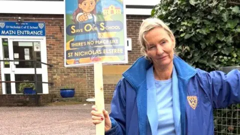 Kate Johnston-Grant Kate Johnston-Grant with medium-length blonde hair, wearing a blue T-shirt, blue jacket with school crest and beige trousers . She is carrying a sign saying "Save our School" and leaning against a metal gate. The school is visible in the background.