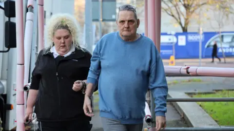 Athena A man handcuffed to a prison officer. He wears a blue jumper and has glasses on his forehead. The prison officer has curly blonde hair and a black top and trousers.