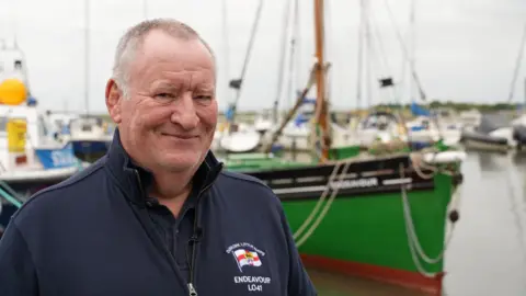 John Fairhall/BBC Cameron McGregor wearing a navy fleece standing on the decking at Leigh Marina, with the green Endeavour boat in the water behind him.