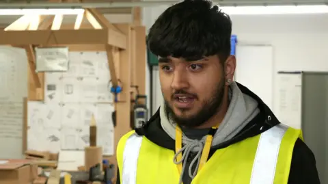 Akeeb Ahmed has black hair and a beard and wears a yellow high-vis jacket over his black hoodie.
