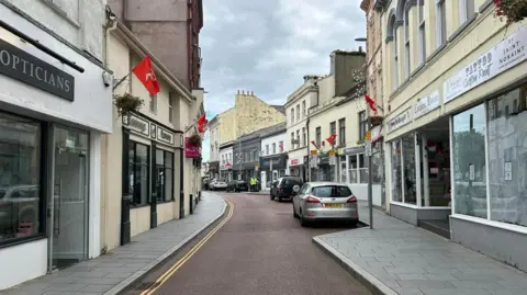 BBC Ramsey high street, with two rows of shops including an opticians, a tattoo parlour and a pharmacy. Red Manx flags are dotted along the street above the shops. The tarmac of the road separating the rows is red, and cars are parked along the right-hand side of the street.