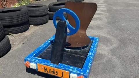 A wooden go-kart with blue wheel and surround, a wooden seat attached to a black base. There is also a number plat which says "kids 1" on it. It is in a school playground, on tarmac and there are black tyres around it. 