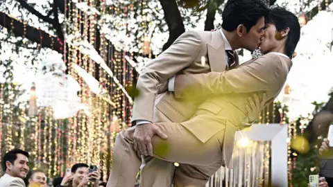 This photo taken on January 10, 2025 shows Thai actors Apiwat "Porsch" Apiwatsayree (L) and Sappanyoo "Arm" Panatkool in beige tuxedos sharing a kiss at their unofficial wedding ceremony in Bangkok. They are surrounded by family and friends taking photos of them under a tree decorated with twinkling lights.