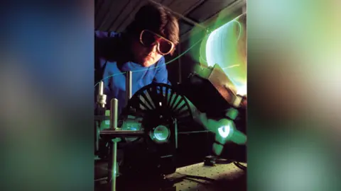 BT Group A photo taken in the 1980s of an engineer working on optical fibre. He wears large protective goggles and a blue jumper. He has brown hair. Fibre wrapped around a cylinder glows a green colour as it is used within a machine.