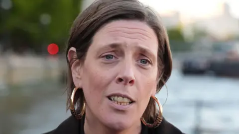 Jess Phillips has short brown hair tucked behind her ears. She is wearing a dark jacket and is forwards.