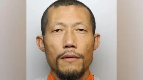 Wiltshire Police Hongchi Xiao looks at the camera with a blank expression in a custody picture. He is balding with dark hair and a dark goatee, and wears a grey sweatshirt over an orange top. 