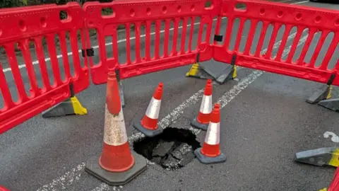 Oxfordshire County Council The sinkhole on the A361. It is surrounded by orange and white road cones and road barriers.